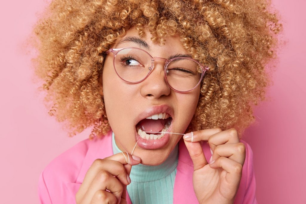 curly-haired woman flossing her teeth with one eye closed