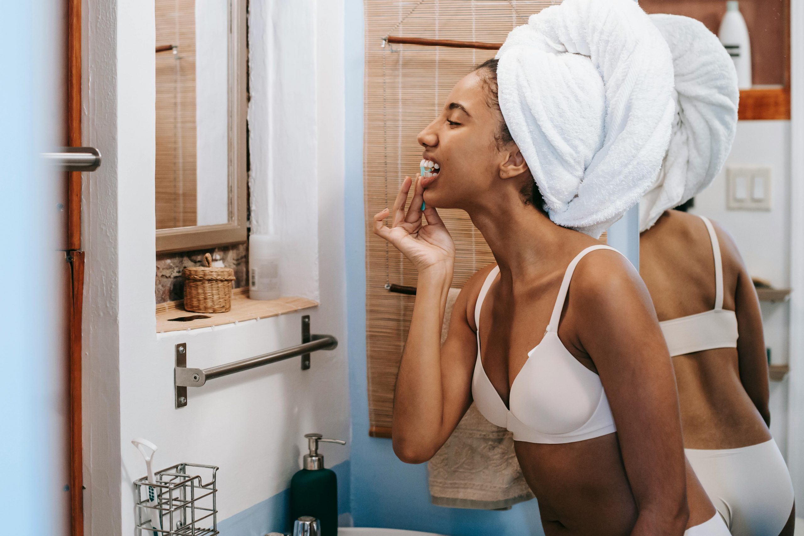 woman with a towel wrapped around her head flossing her teeth in front of a bathroom mirror