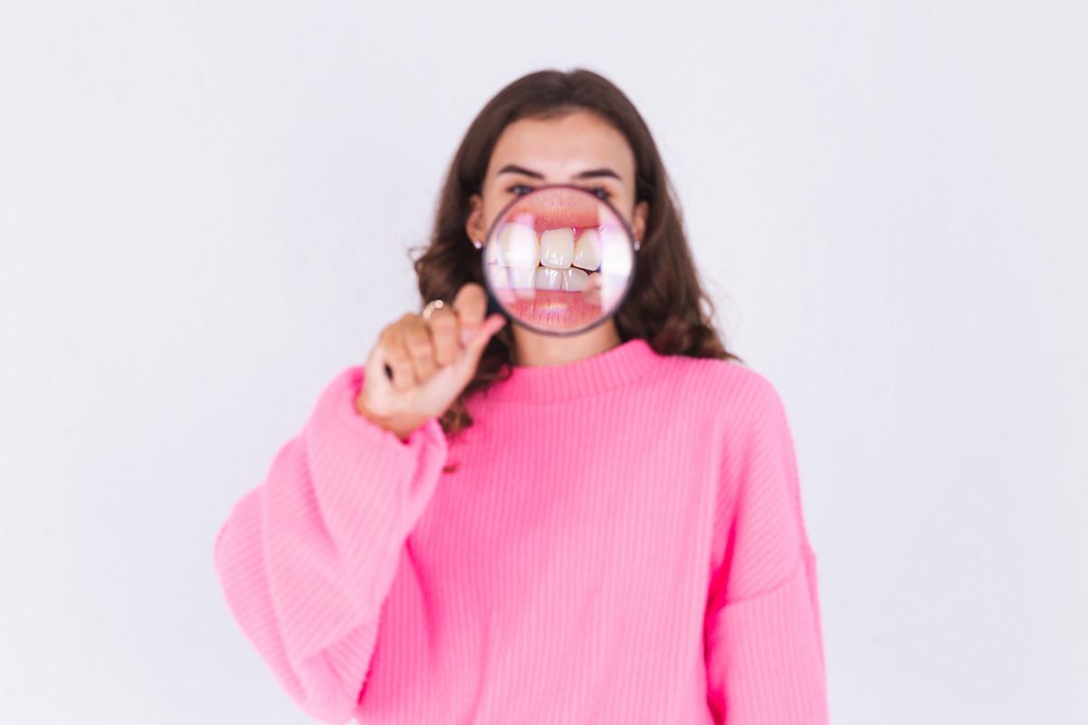 woman in a pink sweater holding a magnifying glass to her mouth, revealing irritated gums
